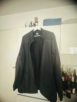 Svart vindjacka från Nike - Snygg svart vindjacka från Nike med diskreta horisontella ränder över axlarna. Jackan har dragkedja framtill, hög krage och är tillverkad i ett lätt, vindtätt material. Perfekt för blåsiga dagar och sportiga outfits.