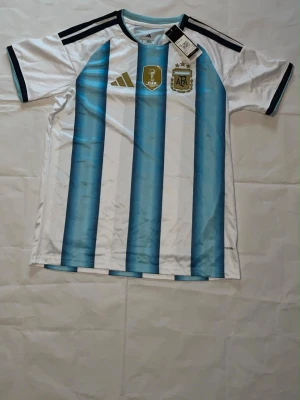 Argentina fotbollströja Adidas storlek L - Säljer en officiell Argentina fotbollströja från Adidas med klassiska blå och vita vertikala ränder. Tröjan har svarta detaljer på axlarna, FIFA- och AFA-emblem på bröstet och är tillverkad i ett lätt, ventilerande material. Perfekt för match eller träning.