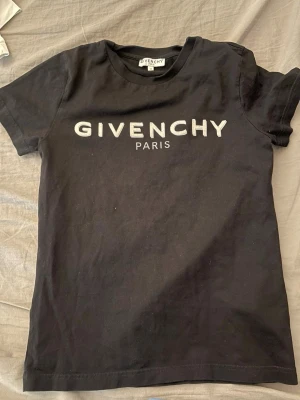 Svart Givenchy t-shirt med logga (barn)  - Snygg svart t-shirt (för barn, 10år?) från Givenchy med stor vit logga och texten 'Paris' på bröstet. Klassisk passform och rund halsringning. Tillverkad i mjuk bomull som känns skön mot huden. Perfekt för dig som vill ha en stilren och trendig look.