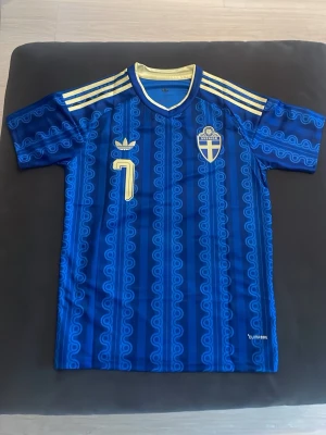 Blå Sverige fotbollströja Adidas - Säljer en blå fotbollströja från Adidas med Sveriges landslagsemblem och nummer 7 i gult. Tröjan har gul krage, tre gula ränder på axlarna och ett unikt mönster i blå nyanser. Perfekt för dig som vill stötta Sverige på match eller träning.