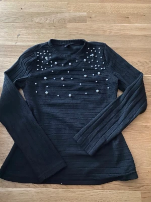 Svart stickad tröja med pärlor SHEIN - Trendig svart stickad tröja från SHEIN i storlek S. Tröjan har långa ärmar och är dekorerad med vita pärlor över bröstet för en snygg detalj. Materialet är mjukt och ribbstickat, perfekt för dig som gillar en stilren men ändå unik look.