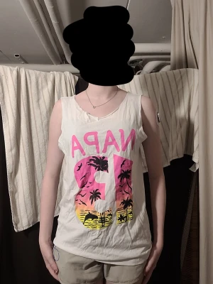 Vit ärmlös topp med tryck - Säljer en vit ärmlös topp med färgglatt tryck framtill och texten 'NAPA 13' i rosa och gult med palmmotiv. På baksidan står det 'I'M IN NAPA BITCH!' i svart och rosa. Toppen har avslappnad passform och rundad halsringning. Passar både S-M-L