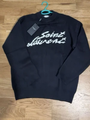 Svart stickad Saint Laurent tröja - Svart stickad tröja från Saint Laurent med vit logotyp i grafisk stil på bröstet. Tröjan har rund halsringning, långa ärmar och ribbade muddar vid ärmslut och nederkant. Perfekt för dig som gillar en clean men ändå statement look.