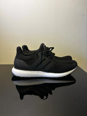 Svarta Adidas Ultra Boost sneakers - Säljer ett par svarta Adidas Ultra Boost sneakers med stickad ovandel och vita Boost-sulor. Skorna har klassiska tre ränder på sidan, svart hälkappa och slitstark yttersula med mönster för bra grepp. Perfekta för dig som gillar sportig och stilren look.