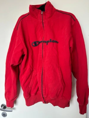 Röd zip hoodie från Champion - Röd hoodie från Champion med dragkedja framtill och broderad logga över bröstet. Tröjan har två fickor och hög krage. 
