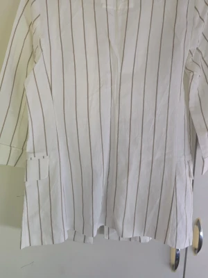 Vit kavaj med smala ränder - Snygg vit kavaj med tunna vertikala beige ränder. Kavajen har klassisk krage, två fickor med lock och trekvartsärmar. Materialet ser ut att vara lätt och luftigt, perfekt för vår och sommar. Enkel och stilren design som passar till många outfits.