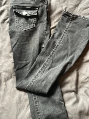 Svarta jeans från True Religion - Säljer ett par svarta jeans från True Religion med bootcut och snygga kontrastsömmar. Jeansen har två bakfickor med lock och dekorativa knappar samt tydliga vita sömmar som ger en cool look. Perfekta för dig som gillar streetwear och vill sticka ut.