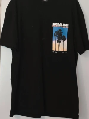 Svart Miami t-shirt med tryck Miami - Cool svart t-shirt från YSMN med färgglatt Miami-tryck fram och bak. Motivet visar palmer mot en solnedgång i blått, rosa och orange. T-shirten har normal passform och korta ärmar, perfekt för en avslappnad streetstyle-look.