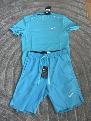 Nike ljusblå träningsset herr - Säljer ett ljusblått träningsset från Nike med t-shirt och shorts. Båda plaggen har Nike-logga i vitt och är tillverkade i ett lätt och ventilerande material, perfekt för träning eller löpning. T-shirten har rund hals och shortsen har dragsko i midjan.