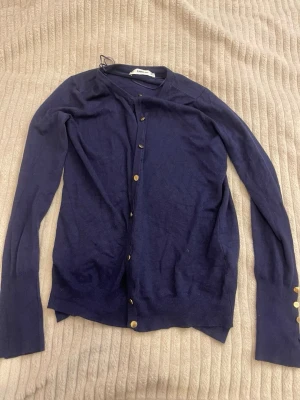 Mörk/marinblå cardigan med guldfärgade knappar - Snygg mörk/marinblå cardigan från ZARA med guldfärgade knappar framtill och på ärmsluten. Tröjan har rund halsringning och är ribbstickad vid muddar och nederkant. Perfekt att slänga över en t-shirt eller linne eller ha det som det är om man vill. 