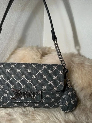 Juicy Couture monogram handväska - Snygg svart och grå handväska från Juicy Couture med JC-monogram över hela väskan. Väskan har en axelrem i svart läder och silverfärgad kedjedetalj samt ett matchande litet myntfodral. Materialet är en mix av textil och konstläder.