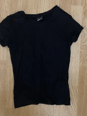 Svart t-shirt från Gina Tricot - En basic svart t-shirt från Gina Tricot i mjuk bomull. Den är figurnära. Modellen är klassisk med rund halsringning och korta ärmar. Perfekt att styla till jeans eller kjol för en enkel och clean look.