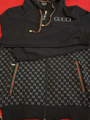 Svart Gucci hoodie med logomönster - Säljer en svart hoodie från Gucci med dragkedja, huva och gröna/röda detaljer. Framsidan har Guccis logomönster och två fickor med färgade kanter. Stora GUCCI-trycket på bröstet och exklusiv metall-dragkedja. Tillverkad i Italien.