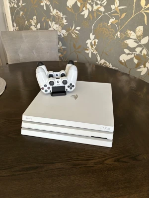 Vit Sony PS4 Pro med två kontroller - Säljer en vit PlayStation 4 Pro med två matchande DualShock 4-kontroller. Konsolen har en stilren design med PlayStation-loggan i silver och kommer med strömkabel och laddstation för handkontroller. Perfekt för gaming och filmkvällar. Tyvärr är den ena kontrollen lite dålig och skulle behöva ett batteri (se sista bild) pris går sjävklart att diskutera 