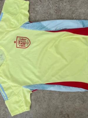 Gul Spanien fotbollströja Adidas - Snygg gul fotbollströja från Adidas med Spaniens landslagsemblem på bröstet. Tröjan har röda och ljusblå detaljer längs sidorna och axlarna samt en röd stjärna ovanför emblemet. Tillverkad i lätt och ventilerande material, perfekt för träning eller match.