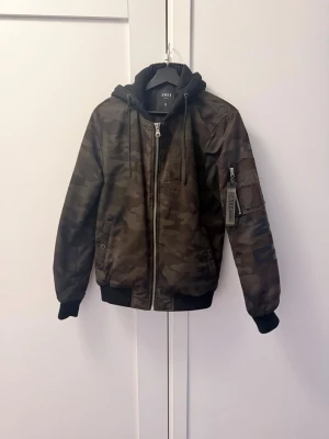 Kamo bomberjacka med huva - Snygg bomberjacka i camouflage-mönster med svart huva och ribbade muddar. Jackan har dragkedja framtill, fickor med tryckknappar och en ärmficka med zip och tag. Perfekt för dig som gillar streetwear och vill sticka ut.