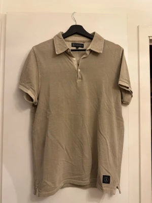 Beige pikétröja från Lagency - Snygg beige pikétröja från Lagency med klassisk krage och knappar framtill. Tillverkad i mjuk bomull och har korta ärmar samt en liten logotyp-patch nedtill. Perfekt till jeans eller shorts för en clean look.