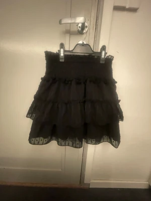 Svart volangkjol - Säljer en svart kortkjol med flera lager volanger och meshdetaljer längst ner. Kjolen har en smockad midja som ger en bekväm passform och ett skirt, luftigt tyg. Perfekt för dig som gillar en söt och trendig stil