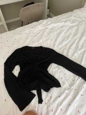 Svart v ringad med knytband - Snygg svart omlottblus från Gina Tricot med långa ärmar och knytband bak. Blusen har v-ringning och är tillverkad i ett mjukt, lätt strukturerat tyg som ger en skön passform. Perfekt att styla med jeans eller kjol.