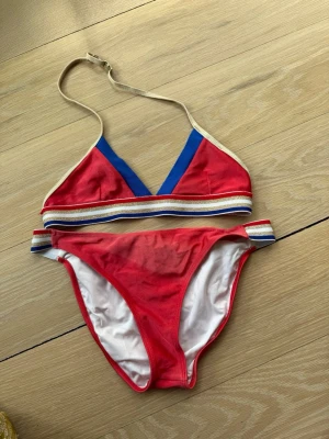 Marc Jacobs Röd och blå bikini med ränder - Snygg bikini i rött och blått med vita och blå ränder på både överdel och underdel. Triangelformad topp med justerbara axelband och sportig känsla. Perfekt för stranden eller poolen i sommar!
