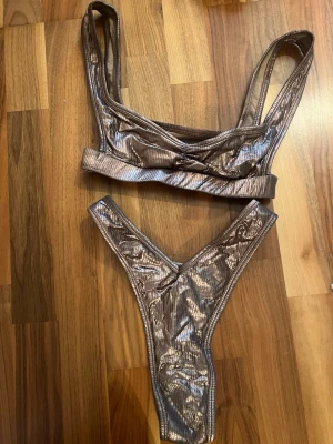 Guldig bikini Oh Polly! - Snygg bikini i glittrigt brunt material med ribbad struktur. Överdelen har breda axelband och en sportig form, medan underdelen är högt skuren med v-formad midja. Perfekt för dig som vill sticka ut på stranden! Från Oh Polly, uppsydd underdel (var alldeles för lång innan, går dock att sprätta upp). Överdel storlek S, underdel storlek M (passar mig med S).