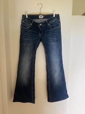 Blå bootcut jeans - Säljer ett par blå bootcut jeans med snygga slitningar och lätt tvättad look. Jeansen har en väldigt låg midja. Perfekta för dig som gillar en avslappnad men trendig stil.