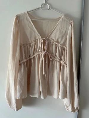 Beige blus med knytband och v-ringning - Söt beige blus med v-ringning och dekorativa knytband framtill. Blusen har volangdetaljer över bysten och är tillverkad i ett tunt, mjukt tyg som ger ett luftigt intryck. Långa ärmar med resår vid ärmslut för en avslappnad look.