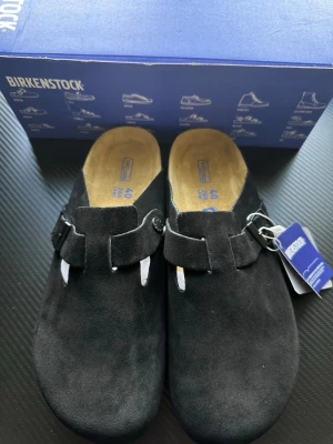 Svarta Birkenstock tofflor i mocka - Säljer ett par svarta Birkenstock tofflor i mjuk mocka med justerbart spänne och klassisk korksula. Tofflorna har öppen häl och bekväm innersula i mocka. Perfekta för dig som vill ha både stil och komfort.