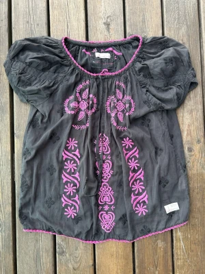 Odd Molly Blus Storlek S/M Svart och Lila - Unik svart blus med puffiga korta ärmar och detaljerad rosa brodyr framtill. Blusen har rund halsringning med rosa kant och ett bohemiskt mönster i brodyren. Tillverkad i ett tunt, luftigt tyg som passar perfekt till jeans eller kjol.