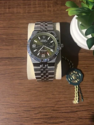 Klocka - Snygg klocka i silver med klassiskt länkarmband och räfflad bezel. Urtavlan är mörk med vita detaljer och tydliga siffror vid 3, 6 och 9. Klockan har datumvisning. Perfekt accessoar för dig som gillar stilrena detaljer. Helt ny, inga skador alls och fungerar perfekt. Priset kan diskuteras!