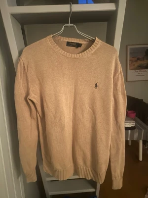 Beige stickad tröja från Polo Ralph Lauren - Klassisk beige stickad tröja från Polo Ralph Lauren med rund halsringning och diskret broderad logga på bröstet. Tröjan har långa ärmar och ribbade muddar vid ärmslut och nederkant. Perfekt för lager på lager och enkel att matcha.