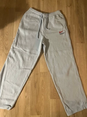 Ljusblåa mjukisbyxor från Nike - Ljusblåa mjukisbyxor från Nike med röd logga och text på vänster lår. Byxorna har resår i midjan med dragsko och raka ben. Tillverkade i mjukt material som känns skönt mot huden. Perfekta för en avslappnad stil.
