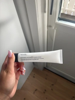 Woods Copenhagen antioxidant handkräm - Restoring Antioxidant Hand Lotion från Woods Copenhagen i en vit tub med svart lock. Handkrämen är vegansk och innehåller antioxidanter som skyddar och återfuktar huden. Perfekt att ha i väskan för mjuka händer hela dagen.