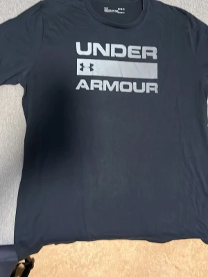 Svart Under Armour t-shirt - Svart t-shirt från Under Armour med stort silverfärgat logotryck på bröstet. Klassisk rund halsringning och korta ärmar. Perfekt för en sportig och avslappnad stil.