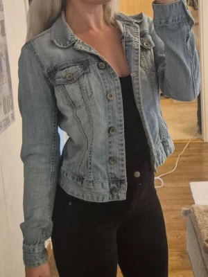 Ljusblå klassisk jeansjacka - Säljer en ljusblå jeansjacka i klassisk modell med knappar framtill och två bröstfickor med lock. Jackan har en figurnära passform och långa ärmar. Perfekt att slänga över en t-shirt eller hoodie för en avslappnad look.