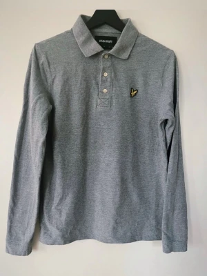  Lyle & Scott pike’ stl S - Stilren långärmad pikétröja från Lyle & Scott i grått. Tröjan har en klassisk krage och knäppning framtill. Den ikoniska örnlogotypen är broderad på bröstet, vilket ger en igenkännbar detalj. Passar utmärkt för en avslappnad men ändå välklädd look. Tröjan är i nyskick. Djur och rökfritt hem.