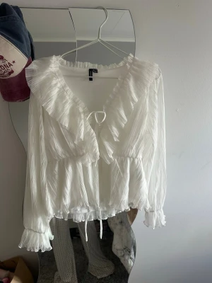 Vit volangblus med knytband - Säljer en romantisk vit blus från vero moda med volangkantad V-ringning och knytband framtill. Blusen har långärmade ärmar med resår och volang vid ärmslut. Tillverkad i ett lätt, plisserat tyg som ger en luftig känsla.