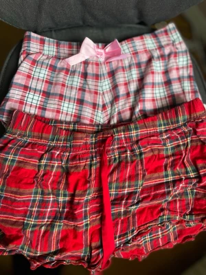 Två rutiga pyjamasshorts med rosett - Säljer två par pyjamasshorts i mjuk bomull. Ena paret är röd- och grönrutig med volangkant och röd dragsko, andra paret är rosa- och vitrutig med söt rosa rosett framtill och spetskant. Perfekta för mysiga kvällar hemma.
