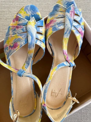 Blommiga pumps med korkklack  - Färgglada pumps med blommigt mönster i blått, gult och rosa. Skorna har öppen tå, rem över foten och justerbart spänne vid ankeln. Klacken är i korkmaterial och ger en somrig känsla. Perfekta för dig som vill sticka ut med din stil.
