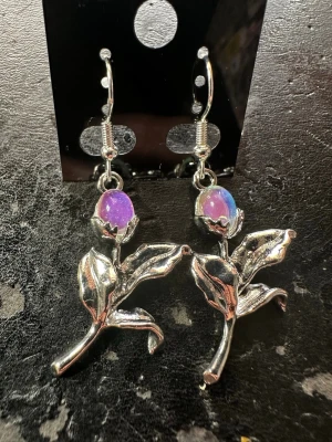 Silvriga örhängen med månstenfärgade pärlor - Unika örhängen i silverfärgad metall formade som blommor med detaljerade blad och stjälkar. Varje blomma har en skimrande månstensfärgad pärla som krona, vilket ger en magisk och färgstark touch. Perfekta för dig som vill sticka ut med coola accessoarer.