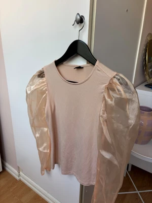 Beige topp med puffiga organzaärmar - Trendig beige topp med rund halsringning och långa, puffiga ärmar i transparent organza. Kroppen är i mjukt tyg medan ärmarna ger en cool kontrast och extra volym. Perfekt för dig som vill sticka ut med en unik detalj på din outfit.