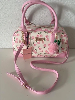Juicy Couture rosa körsbär handväska - Supergullig handväska från Juicy Couture i vitt och ljusrosa med körsbärsmönster. Väskan har både handtag och axelrem, guldfärgade detaljer och en fluffig rosa nyckelring. Perfekt för dig som gillar söta accessoarer och vill sticka ut.