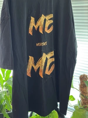 Svart t-shirt med ME VS ME-tryck - Cool svart t-shirt med stort gult och orange tryck på framsidan där det står 'ME versus ME'. T-shirten har korta ärmar och en avslappnad passform. Perfekt för dig som gillar statement-plagg och streetwear vibes.