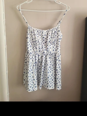 Vit blommig ärmlös sommarklänning - Söt vit kortklänning med blått blommönster och smala axelband. Klänningen har knappar framtill och en lätt rynkad midja som ger en avslappnad passform. Perfekt för varma dagar och har en fräsch, somrig vibe.