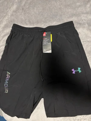 Svarta shorts från Under Armour M - Snygga svarta shorts från Under Armour med elastisk midja och dragsko. Loggan och texten 'ARMOUR' i regnbågsfärgad stil på benet. Tillverkade i lätt och snabbtorkande material, perfekta för träning eller chill.