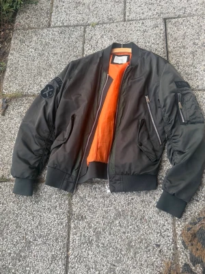 Svart bomberjacka med patchar - Säljer en svart bomberjacka med orange innerfoder och coola patchar på ärmen, bland annat med texten 'FREQUENCY' och en rund logga. Jackan har dragkedja, fickor med lock och klassisk ärmficka med dragkedja. Tillverkad i glansigt syntetmaterial.