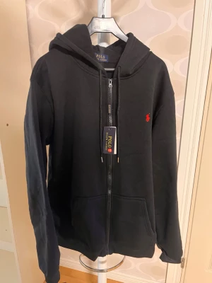 Svart tunn kofta  från Polo Ralph Lauren - Snygg svart tunn kofta  med dragkedja från Polo Ralph Lauren. Klassisk design med huva, fickor framtill och den ikoniska röda loggan broderad på bröstet. Perfekt för en chill och stilren look. Mjuk bomullskvalitet och sköna muddar vid ärmar och midja. STRL XXL