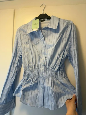 Blå randig skjorta från Gina Tricot - Snygg blå skjorta från Gina Tricot med tunna vertikala ränder och markerad midja med resår. Skjortan har klassisk krage, knappar framtill och långa ärmar. Tillverkad i 50% certifierad bomull. Perfekt för en trendig och fräsch look.