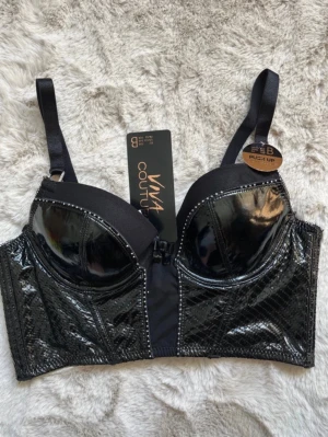 Svart push-up bh från Viva Couture - Snygg svart push-up bh från Viva Couture med glansigt material och dekorativa sömmar. Bh:n har justerbara axelband och en lång modell med breda sidor. Framsidan har en dragkedja och kuporna är förstärkta för extra lyft.
