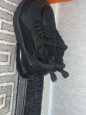 Nike Air Max 270 helsvarta sneakers - Snygga helsvarta Nike Air Max 270 sneakers med stor synlig Air-enhet i hälen. Ovandelen är i mesh och syntetmaterial för bra andningsförmåga och komfort. Klassisk Nike-logga på sidan och dragflik bak för enkel påtagning. Perfekta för en streetwear-look.